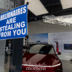 A blue and white sign reading "Billionaires Are Stealing From You" is taped to the glass window of the Tesla Showroom at 860 Washington Street, NYC, during the Picket Tesla protest on March 1, 2025. Through the window, a red Tesla vehicle is visible inside the showroom, while a police officer is seen walking in the background. The reflection in the glass captures the surrounding urban environment, including the High Line pedestrian bridge above the street. This protest, organized by Rise and Resist NYC (@riseandresistnyc), was part of a demonstration against Elon Musk’s political influence and corporate power.