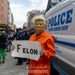 A protester dressed in an orange prison jumpsuit and a Donald Trump mask stands in front of an NYPD Strategic Response Group police van at the Picket Tesla protest at 860 Washington Street, NYC, on March 1, 2025. They hold a white sign with the bold word "FELON" written in black letters, referencing Trump’s legal troubles. The background includes a crowd of protesters holding anti-Musk and anti-Trump signs, as well as NYPD officers monitoring the demonstration. Some of the visible signs in the background read "MUSK WTF?" and "Clear Right Now". The protest, organized by Rise and Resist NYC (@riseandresistnyc), targeted Elon Musk’s influence and corporate power, drawing connections between billionaires, political corruption, and law enforcement.