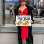 A protester dressed in a satirical wealthy elite costume stands outside the Tesla Showroom at 860 Washington Street, NYC, during the Picket Tesla protest on March 1, 2025. She wears a lavish red dress, a black fur coat, fishnet stockings, and gold gloves, accessorized with pearl necklaces, earrings, and a black top hat adorned with red sequins. Holding a sign that reads "TRILLIONAIRES for TRUMP" with an illustration of stacked gold coins, she embodies a mock portrayal of the ultra-rich supporting Trump and Musk.
Behind her, a blue sign reading "BILLIONAIRES ARE STEALING FROM YOU" is taped to the Tesla showroom window, reinforcing the protest’s message against corporate greed and wealth inequality. A Tesla Optimus robot display and reflections of protesters are visible through the glass, emphasizing the contrast between luxury technology and activist resistance. The protest, organized by Rise and Resist NYC (@riseandresistnyc), drew demonstrators criticizing Musk’s influence, Trump’s policies, and the broader issue of unchecked billionaire power.