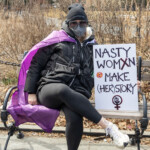 A woman participates in the Women's March held on March 8th, 2025, at Washington Square Park, New York City. She is seated confidently on a park bench, leaning back comfortably while holding a powerful protest sign beside her. The participant is dressed warmly in a black jacket, black leggings, and white sneakers, complemented by a distinctive purple cape that adds vibrant color to her attire. She accessorizes with large, reflective sunglasses and a black knit beanie.
Her sign boldly states:
"NASTY WOMXN MAKE (HER)STORY",
with the word "women" creatively altered to "womxn," a feminist term used to include a broader spectrum of identities. Below the text is a prominent feminist symbol and a decorative sticker.
The photo captures an atmosphere of determined activism amidst a casual setting, highlighting the participant’s strength and expressive support for feminist and women's rights issues. A woman participates in the Women's March held on March 8th, 2025, at Washington Square Park, New York City. She is seated confidently on a park bench, leaning back comfortably while holding a powerful protest sign beside her. The participant is dressed warmly in a black jacket, black leggings, and white sneakers, complemented by a distinctive purple cape that adds vibrant color to her attire. She accessorizes with large, reflective sunglasses and a black knit beanie.