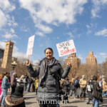 A woman stands powerfully amid the crowd at the Women's March held on March 8th, 2025, at Washington Square Park in New York City. She is holding up two handmade signs, standing out against a vibrant blue sky scattered with birds overhead. She wears a dark grey, puffy winter coat, jeans, and accessorizes with a black-and-white patterned scarf, projecting determination and solidarity.
The sign in her right hand prominently reads:
"SAVE my SiStER",
with "SiStER" highlighted boldly in red letters.
Her left-hand sign carries a sobering message stating:
"7220 WOMEN 13320 CHILDREN DEAD!",
highlighting critical social and humanitarian issues, likely referencing women and children affected by global conflicts or injustices.
The scene captures her determined advocacy amidst fellow demonstrators, amplifying the powerful message of support, awareness, and urgency during this significant gathering.