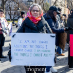 A woman stands solemnly yet assertively holding a protest sign prominently displaying a powerful quote by activist and scholar Angela Davis during the Women's March on March 8th, 2025, at Washington Square Park, New York City. She is dressed warmly in a dark coat, black pants, colorful patterned scarf around her neck, glasses with distinctive frames, and black gloves.
The sign she holds reads clearly and deliberately:
"I AM NO LONGER ACCEPTING THE THINGS I CANNOT CHANGE, I AM CHANGING THE THINGS I CANNOT ACCEPT."
– Angela Davis
She stands amidst a crowd of fellow protesters, united by a shared goal of advocating for social justice, equality, and systemic change.