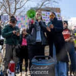 Speaker and activist Evie K passionately addresses the crowd through a green megaphone at the Women's March on March 8th, 2025, at Washington Square Park, New York City. She stands centrally, speaking with conviction and energy. Evie K is dressed comfortably, wearing a black jacket and a grey hoodie, holding a phone with notes or messages in her left hand.
Standing immediately to her left is Evie Joy, attentively supporting the speaker while looking down thoughtfully. Evie Joy wears glasses, a green jacket, dark pants, and has her hair styled up neatly. To the far left of the frame is Jay W. Walker, attentively listening, wearing a dark maroon jacket adorned with various activist buttons and pins, paired with jeans and a hat.
Behind them, protesters hold colorful handmade signs including clearly visible texts:
"I'M WITH HER"
"TRANS RIGHTS ARE HUMAN RIGHTS"
A portable sound system is placed prominently in the foreground, emphasizing the grassroots, community-oriented nature of the event. The atmosphere is powerful, unified, and emotionally charged, focusing on solidarity, social justice, and intersectional activism.