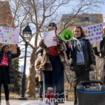 Activist and speaker Kim Russell addresses the energized crowd at the Women's March held on March 8th, 2025, in Washington Square Park, New York City. She passionately speaks from prepared notes, standing confidently at the center of a raised platform alongside other prominent activists. Kim is dressed casually and warmly in a plaid coat over a bright red shirt and blue jeans, holding papers and addressing the audience through a green megaphone.
Activist Evie Joy stands directly next to Kim, assisting with the megaphone and dressed in a black jacket and hoodie. Activist Jay W. Walker is visible at the far left side of the stage, wearing a maroon jacket, checking his phone, and standing attentively.
In the background, two vibrant signs amplify the rally's key messages:
"IF YOU'RE NOT MAD YOU'RE NOT PAYING ATTENTION"
"TRANS RIGHTS ARE HUMAN RIGHTS"
The scene captures a moment of powerful advocacy and collective determination, highlighting intersectional messages addressing women’s rights, trans rights, and broader social justice causes during the Women's March held on March 8th, 2025, in Washington Square Park, NYC.