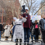 Diana Adams, Esq., a prominent advocate for gender justice and reproductive rights, passionately addresses attendees at the Women's March held on March 8th, 2025, at Washington Square Park in New York City. Diana speaks confidently from prepared notes while activist Jay W. Walker provides vocal amplification through a handheld black megaphone.Surrounding them are fellow activists and demonstrators, including Evie Joy, who stands attentively nearby in a black jacket and hoodie, and another activist dressed in a long black coat and striking yellow-tinted sunglasses. Colorful signs held up by the crowd behind include messages such as "IF YOU'RE NOT MAD YOU'RE NOT PAYING ATTENTION", reflecting the rally's urgency and commitment to social justice, equality, and human rights.