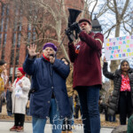 Prominent activist and organizer Leslie Cagan energetically addresses attendees at the Women's March held on March 8th, 2025, in Washington Square Park, New York City. Standing next to her is activist Jay W. Walker, who supports her speech by holding a megaphone. Leslie Cagan passionately engages the crowd, making expressive hand gestures while speaking. She is warmly dressed in a long, dark blue coat, a purple hat, and a bright red scarf, enhancing her vibrant presence at the demonstration.
Jay W. Walker stands confidently beside Leslie, holding the megaphone. He wears a dark maroon jacket adorned with activist pins, dark jeans, sunglasses, and a matching maroon hat. Among the activists behind them is a participant holding a sign reading:
"IF YOU'RE NOT MAD YOU'RE NOT PAYING ATTENTION"
Another sign in the crowd reads:
"MARCH 8TH TODAY IS INTERNATIONAL WOMEN'S DAY...AND IT'S STILL GULF O'CLOCK."
The Women's March gathers activists, advocates, and allies, emphasizing demands for justice, gender equality, reproductive rights, and systemic change, creating an atmosphere of passionate activism and community solidarity.
