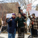 A young activist passionately addresses the crowd through a black megaphone held by Jay W. Walker at the Women's March on March 8th, 2025, at Washington Square Park, NYC. She speaks fervently, embodying youthful determination and courage while standing centrally on a platform. She wears glasses, a dark jacket, and pants, expressing deep conviction through her words and gestures.
Jay W. Walker, dressed in a maroon jacket decorated with activist buttons, glasses, jeans, and a distinctive hat, stands closely behind the young speaker, attentively holding the megaphone for her. Surrounding them are demonstrators carrying vibrant, colorful signs with key messages visible clearly:
"IF YOU'RE NOT MAD YOU'RE NOT PAYING ATTENTION"
"TRANS RIGHTS ARE HUMAN RIGHTS"
"WOMEN OF THE WORLD UNITE"
The rally atmosphere is inclusive and energized, amplifying voices for women's rights, trans rights, and social justice. Nearby, other activists attentively listen and document the speeches, with one participant playing guitar, further enhancing the sense of community and shared purpose.
