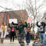 Activist and musician Evie Joy energetically performs a pro-choice anthem at the Women's March on March 8th, 2025, at Washington Square Park, New York City. She enthusiastically sings into a black megaphone, passionately amplifying her voice for reproductive rights and gender equality. Evie Joy wears a dark green jacket, cargo-style pants, and glasses, moving expressively to engage the crowd.
Activist Jay W. Walker is standing supportively behind Evie, wearing a maroon jacket decorated with activist buttons and pins, assisting by holding the megaphone during the performance. Fellow activists join Evie Joy on stage, holding signs with empowering messages clearly visible:
"WOMEN OF THE WORLD UNITE"
"IF YOU'RE NOT MAD YOU'RE NOT PAYING ATTENTION"
"TRANS RIGHTS ARE HUMAN RIGHTS"
A musician to the right side of the frame plays guitar, enhancing the energetic and communal atmosphere. The gathering radiates solidarity, intersectional advocacy, and vibrant grassroots activism.