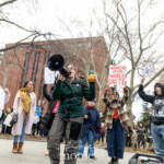 Activist and musician Evie Joy energetically performs a pro-choice anthem at the Women's March on March 8th, 2025, at Washington Square Park, New York City. She enthusiastically sings into a black megaphone, passionately amplifying her voice for reproductive rights and gender equality. Evie Joy wears a dark green jacket, cargo-style pants, and glasses, moving expressively to engage the crowd.
Activist Jay W. Walker is standing supportively behind Evie, wearing a maroon jacket decorated with activist buttons and pins, assisting by holding the megaphone during the performance. Fellow activists join Evie Joy on stage, holding signs with empowering messages clearly visible:
"WOMEN OF THE WORLD UNITE"
"IF YOU'RE NOT MAD YOU'RE NOT PAYING ATTENTION"
"TRANS RIGHTS ARE HUMAN RIGHTS"
A musician to the right side of the frame plays guitar, enhancing the energetic and communal atmosphere. The gathering radiates solidarity, intersectional advocacy, and vibrant grassroots activism.