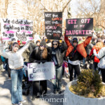 Four activists stand together holding bold and empowering protest signs during the Women's March on March 8th, 2025, at Washington Square Park in New York City. They confidently display their messages advocating women's autonomy, reproductive rights, and social justice.
From left to right:
The first activist, smiling confidently and wearing a light-colored jacket, jeans, sunglasses, and a casual knit cap, holds a large sign reading:
"We didn't start fighting in 1840 to go backward in 2025."
Another participant holds a creatively designed sign featuring three large "E"s, stating:
"EMBOLDENED, EMPOWERED, EMPATHETIC."
The third activist wears sunglasses, a dark jacket, and jeans, holding a strong, clearly written sign reading:
"Not the Church, Not the State, Women shall decide their fate."
The activist on the far right energetically holds up a prominently visible protest sign reading:
"GET OFF MY DAUGHTER."
All demonstrators are dressed appropriately for cold weather, wearing jackets, scarves, and casual attire. Their unified presence highlights collective advocacy, determination, and a passionate demand for reproductive rights, gender equality, and social justice.