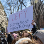 An activist at the Women's March on March 8th, 2025, at Washington Square Park in New York City holds a powerful, clearly visible protest sign with a bold message advocating for defiance and resistance. The handwritten sign prominently displays the word:
"DEFY"
followed by the explanation:
"To Openly Resist or Refuse To Obey."
The participant's hands, clad in dark gloves, firmly grip the sign above the crowd. The demonstrators surrounding the activist are warmly dressed in winter coats, hats, and scarves, showing unity and determination in their collective stance for social justice, women's rights, and activism. Another participant in the background is seen taking photos, capturing the energized atmosphere of the protest.