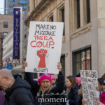 Demonstrators march through the streets near University Place during the Women's March on March 8th, 2025, in New York City. At the forefront, an activist wearing a vibrant pink knitted hat and a dark coat passionately holds a prominently visible protest sign stating:
"MAKE NO MISTAKE THIS IS A COUP. RESIST!"
The sign includes a bold illustration of a raised fist gripping a voting ballot, symbolizing resistance against threats to democracy and political freedom.
Nearby, another participant displays a critical, handwritten message that reads:
"THE BIG BAD MAN AND HIS BIG BAD CLAN THEIR HANDS ARE STAINED WITH RED."
The group of protesters is dressed warmly, reflecting determination as they advocate against authoritarianism, injustice, and for the protection of democratic rights.