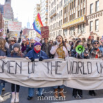 Activists Leslie Cagan, Yanery Cruz, Evie K, and Kim Russell lead a large, determined group of protesters during the Women's March held on March 8th, 2025, marching from Washington Square Park toward Union Square in New York City. Together, they carry a prominent, handmade banner with bold lettering that reads:
"WOMEN OF THE WORLD UNITE!"
Evie K passionately speaks through a green megaphone, rallying the marchers and energizing the crowd, while Leslie Cagan and Yanery Cruz walk steadily and confidently alongside, guiding the demonstration. Kim Russell enthusiastically raises a fist in solidarity, embodying the spirit of activism and unity.
Participants behind the leaders hold numerous vibrant signs, including a rainbow flag symbolizing LGBTQ+ inclusion, and protest placards with feminist messages such as "F*ck the Patriarchy" and "This Country is a Dumpster Fire." The atmosphere is spirited and resolute, with demonstrators dressed warmly in coats, hats, and scarves, marching peacefully yet powerfully through the city streets.