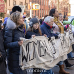 Activists Leslie Cagan, Yanery Cruz, Evie K, and Kim Russell lead a large, determined group of protesters during the Women's March held on March 8th, 2025, marching from Washington Square Park toward Union Square in New York City. Together, they carry a prominent, handmade banner with bold lettering that reads:
"WOMEN OF THE WORLD UNITE!"
Evie K passionately speaks through a green megaphone, rallying the marchers and energizing the crowd, while Leslie Cagan and Yanery Cruz walk steadily and confidently alongside, guiding the demonstration. Kim Russell enthusiastically raises a fist in solidarity, embodying the spirit of activism and unity.
Participants behind the leaders hold numerous vibrant signs, including a rainbow flag symbolizing LGBTQ+ inclusion, and protest placards with feminist messages such as "F*ck the Patriarchy" and "This Country is a Dumpster Fire." The atmosphere is spirited and resolute, with demonstrators dressed warmly in coats, hats, and scarves, marching peacefully yet powerfully through the city streets.