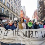 Activists Leslie Cagan, Yanery Cruz, Evie K, and Kim Russell lead a large, determined group of protesters during the Women's March held on March 8th, 2025, marching from Washington Square Park toward Union Square in New York City. Together, they carry a prominent, handmade banner with bold lettering that reads:
"WOMEN OF THE WORLD UNITE!"
Evie K passionately speaks through a green megaphone, rallying the marchers and energizing the crowd, while Leslie Cagan and Yanery Cruz walk steadily and confidently alongside, guiding the demonstration. Kim Russell enthusiastically raises a fist in solidarity, embodying the spirit of activism and unity.
Participants behind the leaders hold numerous vibrant signs, including a rainbow flag symbolizing LGBTQ+ inclusion, and protest placards with feminist messages such as "F*ck the Patriarchy" and "This Country is a Dumpster Fire." The atmosphere is spirited and resolute, with demonstrators dressed warmly in coats, hats, and scarves, marching peacefully yet powerfully through the city streets.
