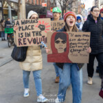A young protester passionately chants slogans as she marches during the Women's March held on March 8th, 2025, moving from Washington Square Park to Union Square in New York City. She enthusiastically shouts while holding two powerful handmade signs. The first sign boldly proclaims, "A WOMAN'S PLACE IS IN THE REVOLUTION," featuring a raised fist graphic symbolizing empowerment. Her second sign presents an impactful quote alongside a painted portrait of activist Malala Yousafzai, stating: "If one man can destroy everything, why can't one girl change it?" Other protesters, warmly dressed in coats, hats, gloves, and scarves, march alongside her, creating a spirited atmosphere of unity, determination, and advocacy.
