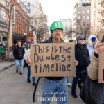 A participant in the Women's March on March 8th, 2025, marching from Washington Square Park to Union Square in New York City, wears a green Luigi hat from the Mario Bros franchise. He carries a cardboard sign humorously yet critically stating, "This is the Dumbest timeline." The expression on his face is serious, complementing the sarcastic tone of the sign. Other demonstrators surround him, moving through the urban backdrop of Manhattan, highlighting a collective mood of frustration and protest.