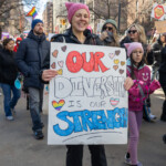 A cheerful protester at the Women's March on March 8th, 2025, walks along Manhattan streets towards Union Square, holding a colorful handmade sign that reads, "Our Diversity is Our Strength," surrounded by decorative hearts and rainbow symbols, emphasizing love and unity. She wears a pink knit hat, representing solidarity, and smiles warmly while marching alongside a young girl wearing a similar pink hat and gloves. Other participants are seen marching in solidarity behind her, reflecting the supportive and inclusive atmosphere of the event.