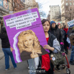 A young protester walks through the streets of Manhattan holding a large purple sign featuring a well-known portrait of Dolly Parton. The sign contains a bold, provocative message addressing former President Trump and his supporters, criticizing their stance on women's rights. The text reads: "Dear Trump and his ass kissers, it’s not our fault you have small dicks, so don’t make it our problem! Sincerely, women everywhere." Behind the protester, a diverse crowd marches forward, holding various signs advocating for women's rights, reproductive justice, and political change. The urban backdrop of New York City, complete with storefronts and apartment buildings, sets the stage for the demonstration, which is part of the larger Women's March on March 8th, 2025.
