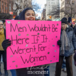A young protester at the Women's March in New York City on March 8, 2025, holds a bright pink sign with bold black lettering that reads, "Men wouldn't be here if it weren't for women." The sign is adorned with small stickers of rainbows, hearts, and stars, adding a playful yet strong statement on gender equality. The protester, dressed in a black winter coat and gloves, smiles while standing among a diverse group of demonstrators marching down a city street. The background features fellow activists, some wearing scarves and hats, with tall buildings and traffic lights framing the scene. The atmosphere is one of solidarity and advocacy for women's rights.