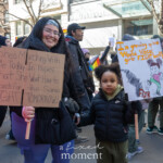 A mother and her young child participate in the Women's March in New York City on March 8, 2025, holding powerful handmade signs. The mother, smiling and wearing a headband and glasses, carries a cardboard sign reading, "She's marching with me today in hopes that she won’t have to march for the same FIGHT tomorrow!" Her child, wearing a black puffer jacket and olive green pants, holds a white sign with a hand-drawn illustration of a girl kicking the word "FEAR" and the message, "I'm growing up in a world where girls FIGHT back!" The scene is filled with protesters walking past storefronts, waving signs and flags, emphasizing the intergenerational fight for gender equality and justice.