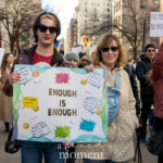 Two protesters stand side by side at the Women's Rights March in New York City on March 8, 2025, holding a colorful sign with the bold statement, "Enough is Enough," surrounded by various handwritten messages advocating for justice, equality, and resistance. One protester, a person in a red top and sunglasses, wears a confident expression, while the other, a woman in a cream quilted jacket and sunglasses, smiles at the camera. The march continues behind them, with numerous demonstrators carrying signs calling for trans rights, racial justice, and resistance against oppression. The crowd is diverse, with individuals of different ages and backgrounds joining together in solidarity for women's and LGBTQ+ rights.