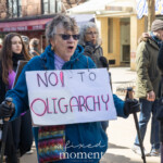 An older woman in a blue fleece jacket and sunglasses marches with determination at the Women's Rights March in New York City on March 8, 2025. She firmly holds a sign that reads "NO! TO OLIGARCHY" in bold, handwritten letters. Using two walking canes, she strides forward, embodying resilience and activism. Around her, fellow protesters chant and hold signs advocating for various causes, including one in the background that reads "Trans Rights Are Women's Rights." The march, filled with passionate voices, highlights a collective call for democracy, equality, and economic justice.