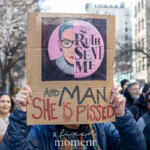 A protester at the Women's Rights March in New York City on March 8, 2025, holds up a cardboard sign featuring an image of Ruth Bader Ginsburg with the words "Ruth Sent Me" on a pink background. Below, the handwritten message reads, "And MAN, SHE IS PISSED," emphasizing the late Supreme Court Justice’s legacy in the fight for gender equality. The demonstrator, wearing a blue puffer jacket and braids, stands amid a crowd of activists and onlookers. Behind them, a diverse group of protesters marches, some carrying signs advocating for women's rights and social justice. The urban setting includes tall buildings and a mix of engaged participants, illustrating the collective energy of the demonstration.