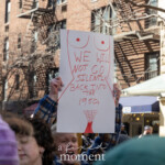 A passionate protester raises a bold sign reading, "We Will Not Go Silently Back Into the 1950s," during the Women's March in New York City on March 8, 2025. The handmade sign features artistic depictions of breasts and a uterus, symbolizing bodily autonomy and reproductive rights. The demonstrator, wearing a blue and black plaid jacket, stands among a diverse crowd of marchers advocating for gender equality and women's rights. In the background, New York City’s urban architecture, including brick buildings with fire escapes, sets the scene for this moment of activism. Other protesters can be seen holding signs and engaging in the demonstration, reinforcing the collective call for progress and resistance against societal regression.