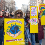 A group of enthusiastic protesters march through the streets of New York City during the Women's March on March 8, 2025, carrying vibrant signs advocating for equality and inclusivity. One prominent sign reads "International Women's Day" with an illustration of hands holding the Earth, symbolizing global unity and women's empowerment. Other signs display messages such as "We Are Equal" and "Love is Love," emphasizing LGBTQ+ rights and gender equality. The demonstrators, dressed warmly in jackets, scarves, and sunglasses, smile and chant as they move forward in solidarity. The urban backdrop of NYC, with its brick buildings and fire escapes, frames this moment of activism and celebration.