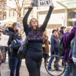 A protester stands tall at the NYC Women's March on March 8, 2025, holding a striking sign that reads "Pussy Grabs Back!" with illustrations of fierce-looking cats. Dressed in black, the woman wears a T-shirt with pink handwritten text that appears to read "Tits & Correct Opinions," adding to her bold statement. She raises her sign high with confidence, embodying the spirit of resistance and empowerment. Around her, other demonstrators hold their own messages advocating for equality and justice. The background captures a lively scene of activists, some wearing masks, as they march through the city streets. A bicycle wheel peeks into the frame, adding to the dynamic urban atmosphere.