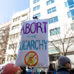 A demonstrator at the NYC Women's March on March 8, 2025, raises a white inflated sign with bold purple and blue lettering that reads "ABORT THE OLIGARCHY." Below the text, a symbol of a crossed-out money bag with a crown emphasizes the anti-wealth concentration message. The sign stands out against the backdrop of modern city buildings, leafless trees, and a crowd of activists marching through the streets. Other protesters are visible holding signs, wearing beanies and sunglasses, braving the crisp weather as they chant and advocate for systemic change.