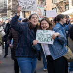 Two young women passionately engage with the crowd at the NYC Women's March on March 8, 2025, holding handmade signs advocating for women's rights. The woman on the left holds a sign that reads "INTERSECTIONAL FEMINISM: ALWAYS" with an illustration of a raised fist, emphasizing the need for inclusivity in the feminist movement. The woman on the right holds a sign that says "STOP CRIMINALIZIN' PREGNANCY!" highlighting the ongoing fight for reproductive justice. Their faces are animated with determination as they march alongside a diverse crowd of demonstrators holding various protest signs in the background. City buildings, fire escapes, and urban architecture frame the scene, underscoring the grassroots energy of the movement.