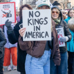 A protester at the NYC Women’s March on March 8, 2025, holds a bold black-and-white sign reading “NO KINGS IN AMERICA,” making a clear statement against authoritarianism and political overreach. Wearing a baseball cap labeled “Dairy Boy,” the individual is dressed casually in a brown jacket and blue jeans, standing among a crowd of demonstrators. Behind them, other protesters hold signs with messages such as "Nobody Elected Musk" and "Women's Rights Are Human Rights." The urban backdrop features tall buildings and fire escapes, framing the demonstration in the heart of New York City. The crowd's engagement and determination reflect the march’s themes of democracy, equality, and resistance.