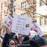A demonstrator at the NYC Women’s March 2025 holds a handwritten sign that reads, “Remember when you voted to take away MY RIGHTS? Yeah, me too!” accompanied by a Progress Pride Flag. The sign is prominently raised above a crowd of activists, who are also holding various protest messages, including one that states “Women Are Human” and another with a crossed-out symbol rejecting voter suppression. The urban backdrop features classic New York City architecture with fire escapes and leafless trees, indicating a winter setting. The atmosphere of the protest is energetic, with many participants expressing strong emotions as they demand equality and justice.