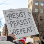 A close-up of a protest sign at the NYC Women's March 2025 reads "RESIST → PERSIST → REPEAT," with a clenched fist symbolizing resistance. The sign is held high amidst a sea of demonstrators carrying various messages, including a partially visible one on the right with bold, colorful letters. The background features New York City's urban landscape with classic brick buildings and leafless trees, indicating a winter setting. The hands holding the sign, clad in gloves and warm clothing, emphasize the cold season. The determined energy of the crowd reflects a strong message of persistence and activism in the fight for women's rights and social justice.