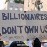 A protester raises a handmade cardboard sign reading "BILLIONAIRES DON'T OWN US" with a handwritten message below urging people to "Join your community. Fight back." The bold black letters stand out against the neutral backdrop of New York City's brick buildings and modern high-rises. The protester, dressed in warm winter clothing with gloves, grips the sign firmly as they march in unison with a diverse crowd of demonstrators advocating for economic justice and corporate accountability. Other protest signs, including one with a rainbow flag, are visible in the background, emphasizing the intersectionality of issues at the rally.