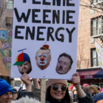 A marcher holds a protest sign that reads "TEENIE WEENIE ENERGY," featuring edited images of well-known political figures, including one with a clown face and another wearing a beanie propeller hat. The sign is aimed at mocking certain politicians and public figures. The crowd surrounding the protester is diverse, filled with individuals carrying colorful banners, signs, and pride flags. Some marchers are smiling and engaging with each other, while others wear winter hats and scarves, braving the cold as they make their way through New York City streets. The march is energetic, with demonstrators voicing their discontent through humor and satire.