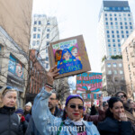 A demonstrator at the NYC Women's March 2025 raises a colorful protest sign that reads, "If the Patriarchy is Heavy, Let's Make it Collapse," accompanied by an illustration of three women of different backgrounds standing together. The protester, wearing a blue denim jacket, sunglasses, a purple headband, and bold red lipstick, looks determined as she participates in the march. Behind her, several other protesters hold signs advocating for trans rights and gender equality, contributing to the powerful atmosphere of solidarity and activism. The crowd is diverse, with people of all ages and identities marching together under a clear blue sky, surrounded by the urban landscape of New York City.