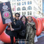 Three women stand together in front of a large red abstract sculpture in New York City, participating in the NYC Women's March 2025. The woman on the left holds a striking protest sign designed like an arcade game interface with the words "SMASH THE PATRIARCHY" along with buttons labeled "RESIST." She wears an all-black outfit, sunglasses, and a collection of feminist pins on her coat. The woman in the middle, dressed in a black coat with a crossbody bag, smiles warmly. The woman on the right, in a bold leopard-print coat with a pink scarf, exudes confidence with her hands on her hip. The background captures the vibrant cityscape with classic NYC architecture, street signs, and other protesters marching for gender equality.