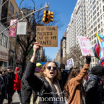A passionate protester raises a bold sign that reads "KEEP YOUR POLITICS OUT OF MY PANTS" during the NYC Women’s March 2025. She wears sunglasses, a black jacket, and has her mouth open, seemingly chanting as she marches forward with the crowd. Surrounding her are numerous demonstrators carrying signs with powerful messages, including "Viva La Vulva" and other advocacy posters, as well as a vibrant trans pride flag waving in the wind. The rally takes place in a bustling cityscape with tall white buildings, traffic lights, and storefronts like Bank of America and food vendors visible in the background. The atmosphere is charged with energy, unity, and determination as people from diverse backgrounds gather to demand reproductive rights, gender equality, and social justice.