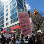 A demonstrator holds up a striking pink sign quoting Ntozake Shange, reading "Where there is a womxn, there is magic." The sign stands tall against a backdrop of modern glass buildings and NYC traffic lights, emphasizing the urban setting of the 2025 Women's March. Other participants in the crowd carry powerful messages, including a fiery sign stating "You Are Right to Fear Our Rage", another with an altered American flag that warns "Wake Up and Smell the Fascism," and various other protest placards. Protesters, dressed warmly in hats and scarves, reflect diverse backgrounds as they march together, chanting and advocating for women's rights, reproductive freedom, and social justice. The energy of the event is palpable, with a strong sense of solidarity and empowerment resonating through the streets.