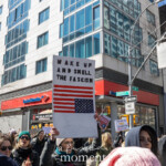 A protester prominently holds up a modified American flag sign with bold black text reading "Wake Up and Smell the Fascism." The flag's stars and stripes appear distorted, emphasizing the message of political urgency and resistance. The demonstrator, clad in a dark jacket, stands among a large, engaged crowd, many holding their own protest signs. In the background, other placards are visible, including one stating "The war on our rights is a war on 70% of us." The setting is an urban environment, with Bank of America, The Halal Kitchen, and street signs for University Place and 14th Street clearly visible, placing the protest in downtown New York City. Protesters display determined expressions, marching for democracy, human rights, and women's rights, with the scene reflecting a powerful moment of civic engagement and activism.