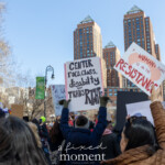 Protesters raise bold signs advocating for intersectional feminism and resistance during the NYC Women's March 2025. One sign prominently states, "CENTER race, class, disability, and TRANS RIGHT," emphasizing inclusivity in the fight for justice. Another sign, held high, reads, "A WOMAN'S PLACE IS IN THE RESISTANCE," written in red and black lettering with a fist graphic, symbolizing strength and defiance. Surrounding the scene, additional signs and trans pride flags wave above the crowd. The protest unfolds in an urban setting with towering buildings and street lamps framing the demonstration. The marchers, dressed warmly, exhibit determination and unity, reinforcing a collective demand for social justice, equality, and trans rights.