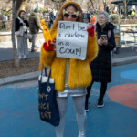 A protester at the NYC Women's March 2025 makes a striking statement both in fashion and activism. Dressed in a bright yellow faux-fur coat, a metallic silver skirt, and knee-high silver platform boots, the marcher holds a sign reading "Don't be a chicken in a coup!" The outfit is accessorized with red gloves, sunglasses, and a fuzzy hood, giving a playful yet assertive presence in the crowd. A tote bag with a visible slogan, “DON’T TOUCH SHIT,” adds to the rebellious energy. The backdrop features a city park with barren winter trees, blue and red painted pavement, and other demonstrators, some observing while others actively participate in the march. This image captures the intersection of humor, political resistance, and bold self-expression at the event.