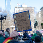 A demonstrator at the NYC Women's March 2025 holds a cardboard sign with the acronym "DOGE" cleverly redefined as "Deranged Oligarchs are Grifting Everything." The sign, outlined in blue, critiques the influence of the wealthy elite on politics and society. The person holding it is seen from behind, wearing a tan beret and a green jacket, with a rainbow pride flag draped over their shoulders. Surrounding them, a crowd of protesters holds various signs, including one in the background reading "Feminism Not Fascism." The location is identifiable as Union Square Park in New York City, with its recognizable equestrian statue and historic buildings in the backdrop. The atmosphere is charged with energy as activists raise their voices for justice and equality.