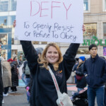 A smiling protester raises a sign that reads "DEFY – To Openly Resist or Refuse to Obey" in bold red and black letters during the NYC Women's March 2025 in Union Square, New York City. She wears a black hoodie, light blue jeans, and carries a white crossbody bag as she stands among a diverse crowd of demonstrators advocating for women's rights and social justice. Other marchers, dressed in warm clothing, hold up additional protest signs. Behind them, a Whole Foods store and retail buildings create an urban backdrop, with pedestrians and fellow activists filling the streets. The event embodies the spirit of resistance and empowerment, urging action against oppressive systems.