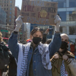 A demonstrator raises a handmade sign that reads "The Power of the People is Stronger Than the People in Power" in bold mixed-color lettering at the NYC Women’s March 2025 in Union Square, New York City. The protester wears a black face mask, a denim jacket with a keffiyeh scarf draped over their shoulders, white gloves, and beige pants. Behind them, another marcher holds a large black poster with images of activists and the phrase "They weren’t all for nothing." The scene is filled with other demonstrators, some wearing backpacks and winter clothing, as they march through the city streets. The background features towering glass buildings, retail shops, and a police department logo visible on a sign. The sunlight creates a dramatic effect, shining down on the protest, symbolizing resilience and collective action.