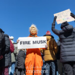 At the NYC Women’s March 2025 in Union Square, New York City, artist and activist @mch463 (Mike Hisey) stands in the crowd wearing an orange jumpsuit and a Donald Trump mask, holding a sign that reads "FIRE MUSK." The costume, symbolizing incarceration, makes a strong political statement about accountability and corporate power. In the foreground, another protester, dressed in a black puffer jacket and gloves, raises a sign that reads "GRAB 'EM BY THE MIDTERMS," a play on Trump's infamous quote, linking it to electoral action. The march is filled with demonstrators in winter attire, standing under a bright blue sky, with the W Union Square hotel visible in the background. The energy of the crowd reflects a collective demand for justice, reform, and resistance against corporate and political figures.