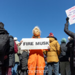 At the NYC Women’s March 2025 in Union Square, New York City, artist and activist @mch463 (Mike Hisey) stands in the crowd wearing an orange jumpsuit and a Donald Trump mask, holding a sign that reads "FIRE MUSK." The costume, symbolizing incarceration, makes a strong political statement about accountability and corporate power. In the foreground, another protester, dressed in a black puffer jacket and gloves, raises a sign that reads "GRAB 'EM BY THE MIDTERMS," a play on Trump's infamous quote, linking it to electoral action. The march is filled with demonstrators in winter attire, standing under a bright blue sky, with the W Union Square hotel visible in the background. The energy of the crowd reflects a collective demand for justice, reform, and resistance against corporate and political figures.